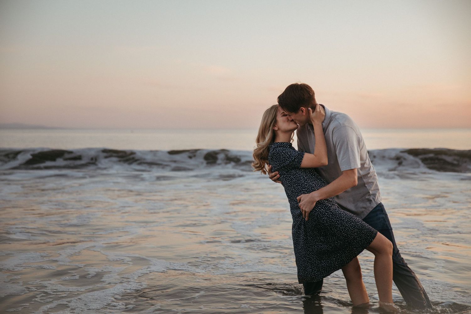 A romantic couple embraces while standing in ocean waves during golden hour sunset.