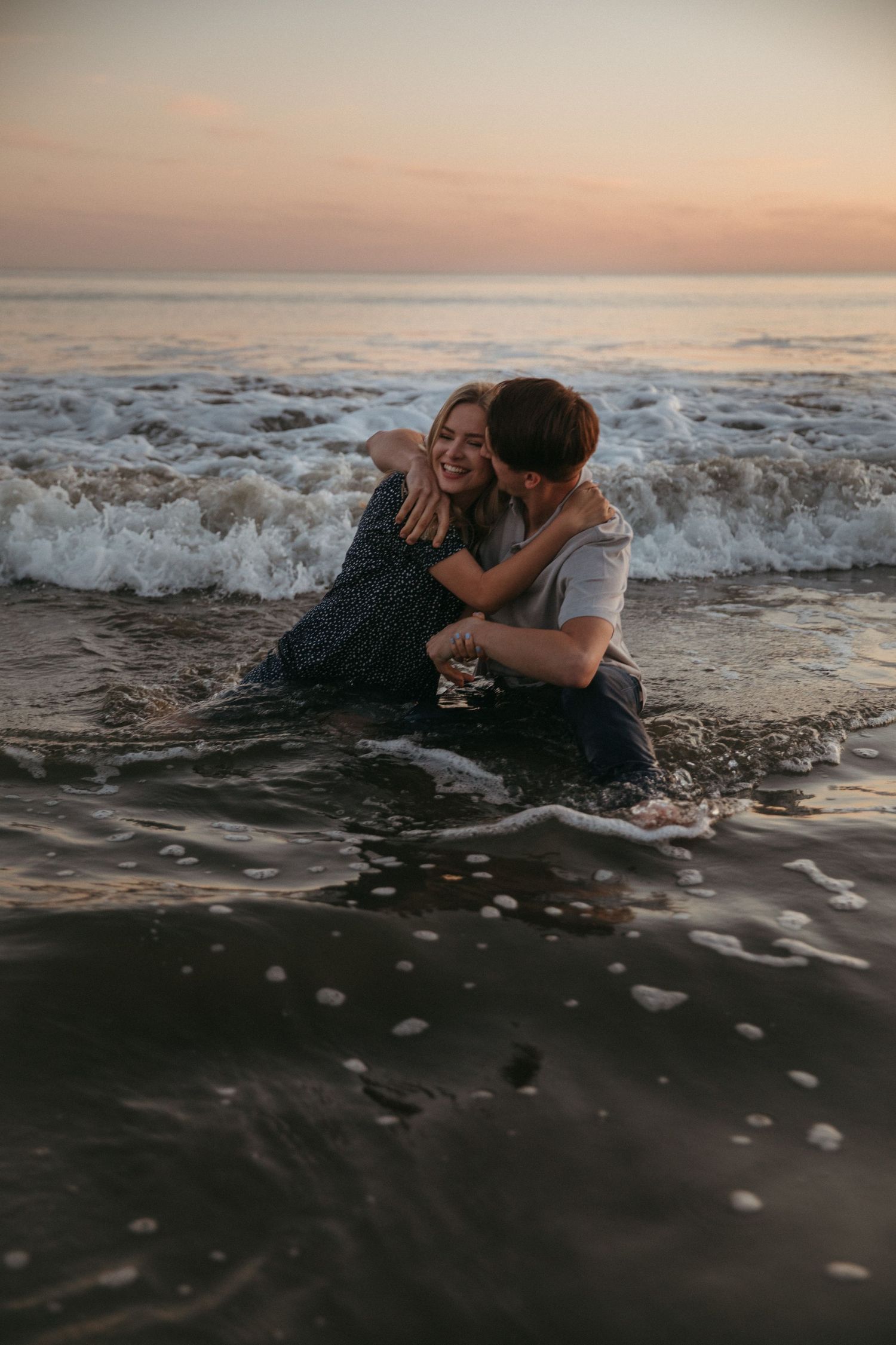 A couple laughs together while sitting in crashing ocean waves during sunset.