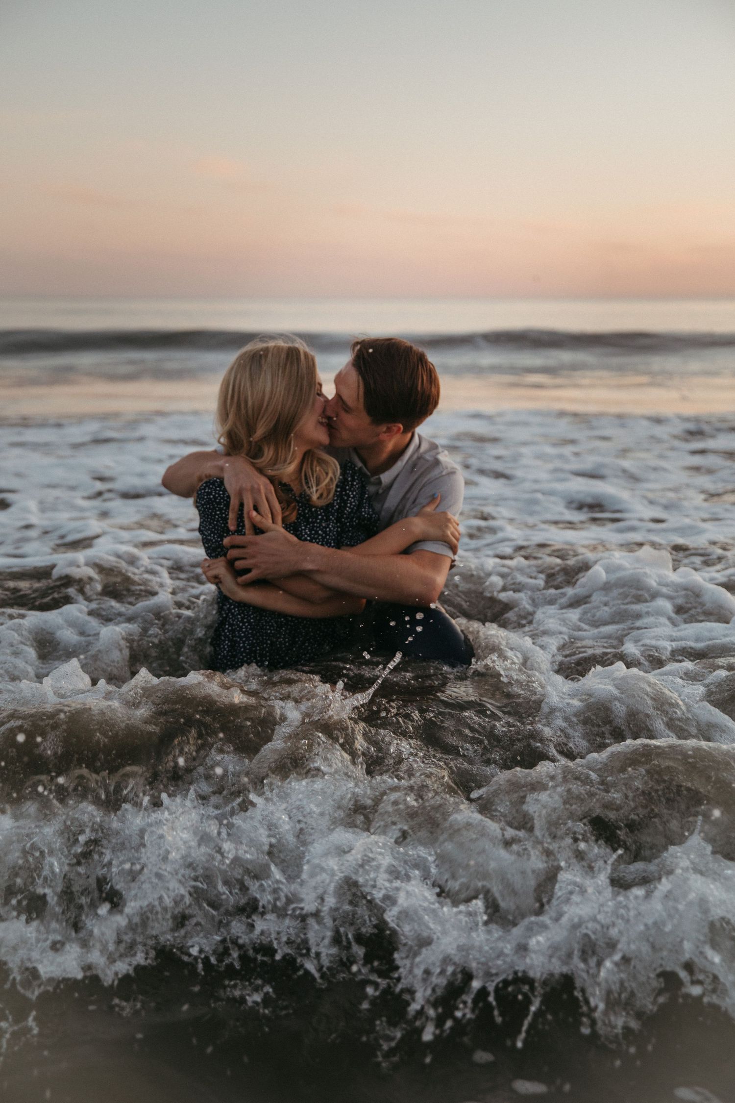 Two people embrace in ocean surf as waves crash around them at dusk.