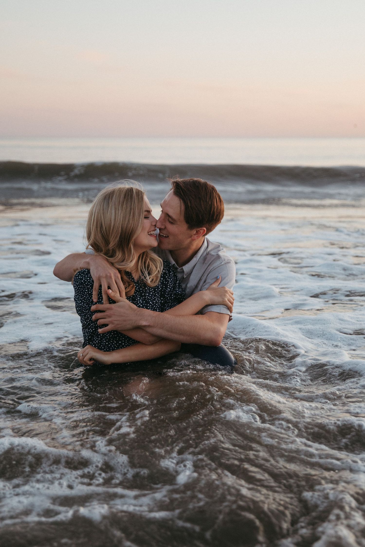 A romantic couple holds each other close while sitting in gentle ocean waves at sunset.