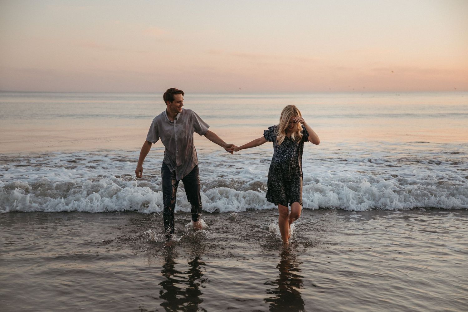 A couple holds hands while walking through shallow ocean water during sunset.