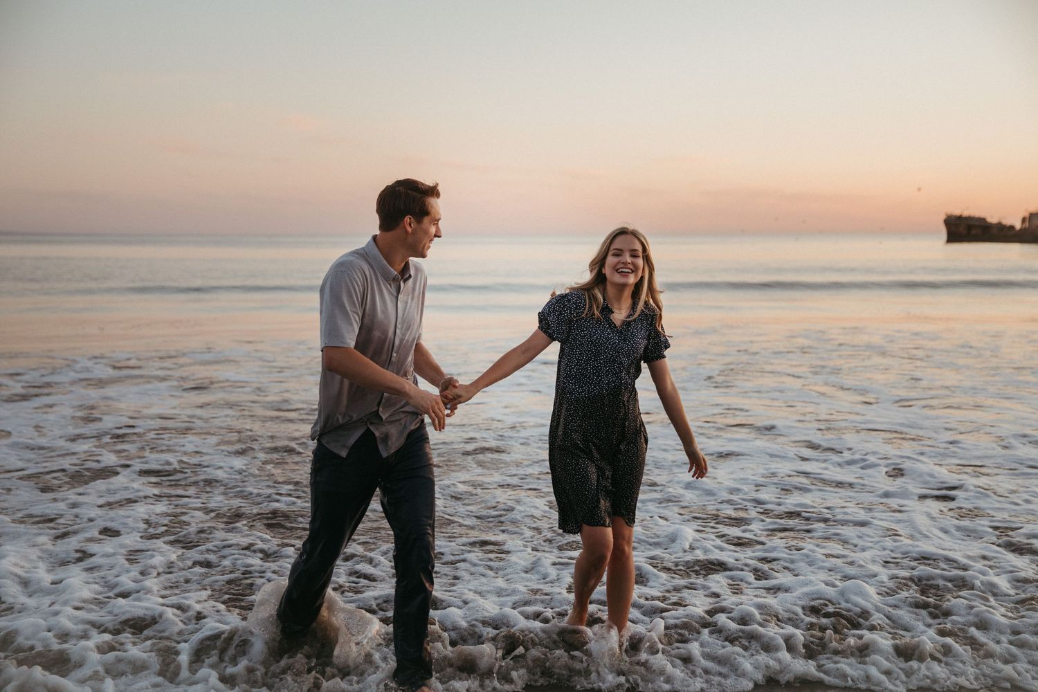 A couple walks hand in hand through ocean waves during golden hour.