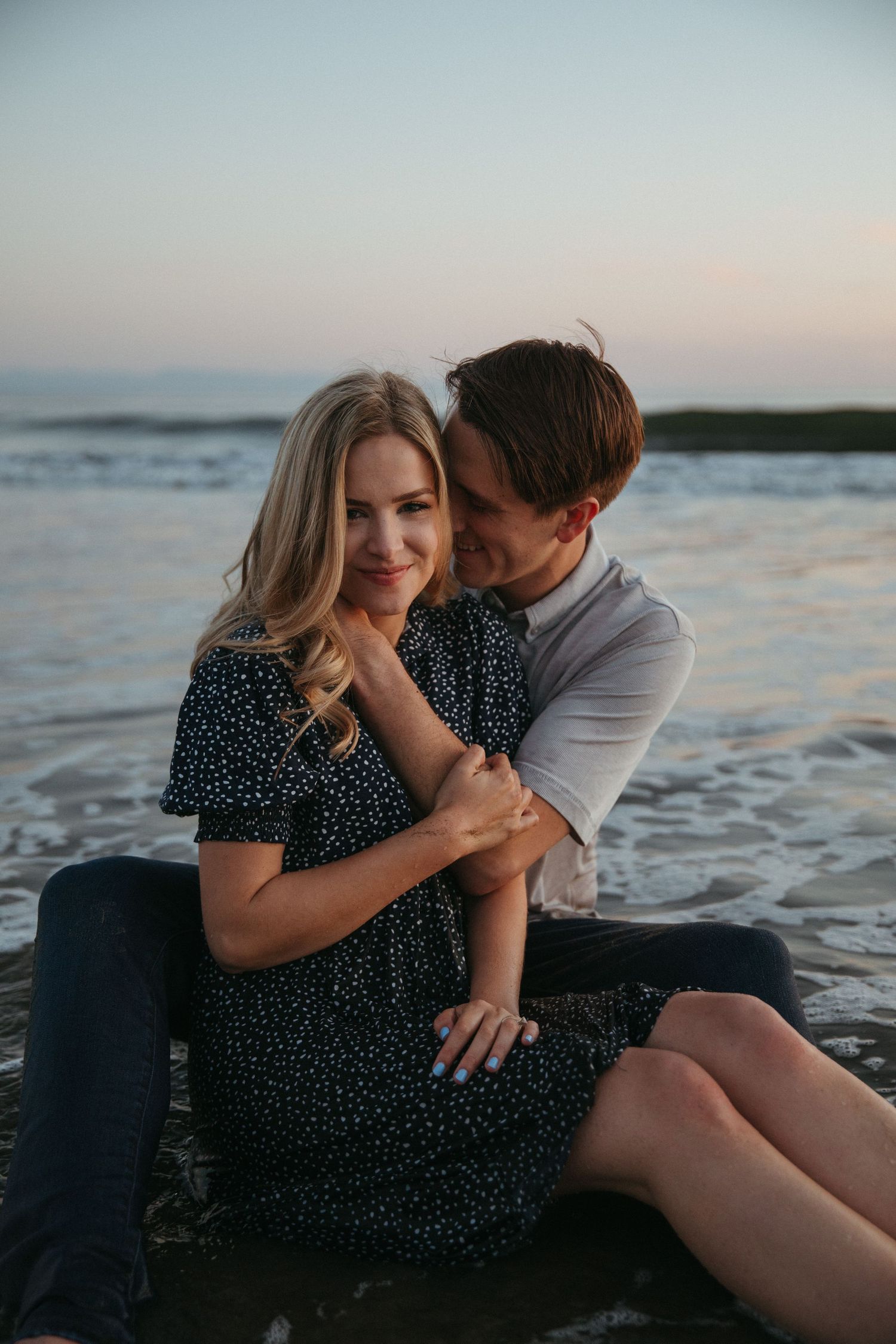 A romantic couple embraces on the beach at sunset, with the woman wearing a polka dot dress.