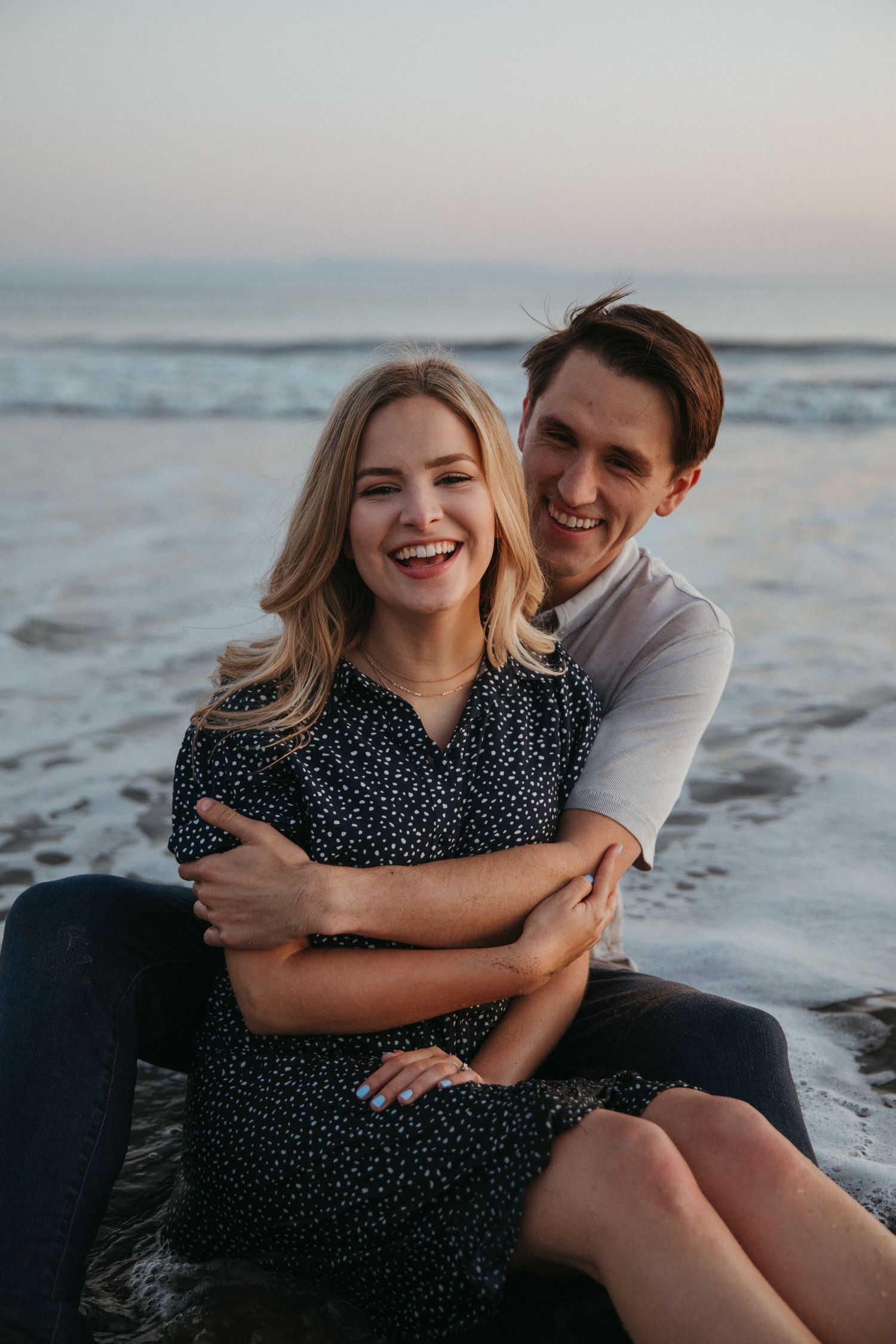 A couple laughs together while sitting on the beach during sunset.