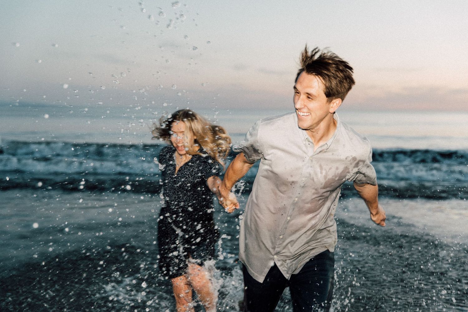 Playful couple running through ocean spray at sunset.