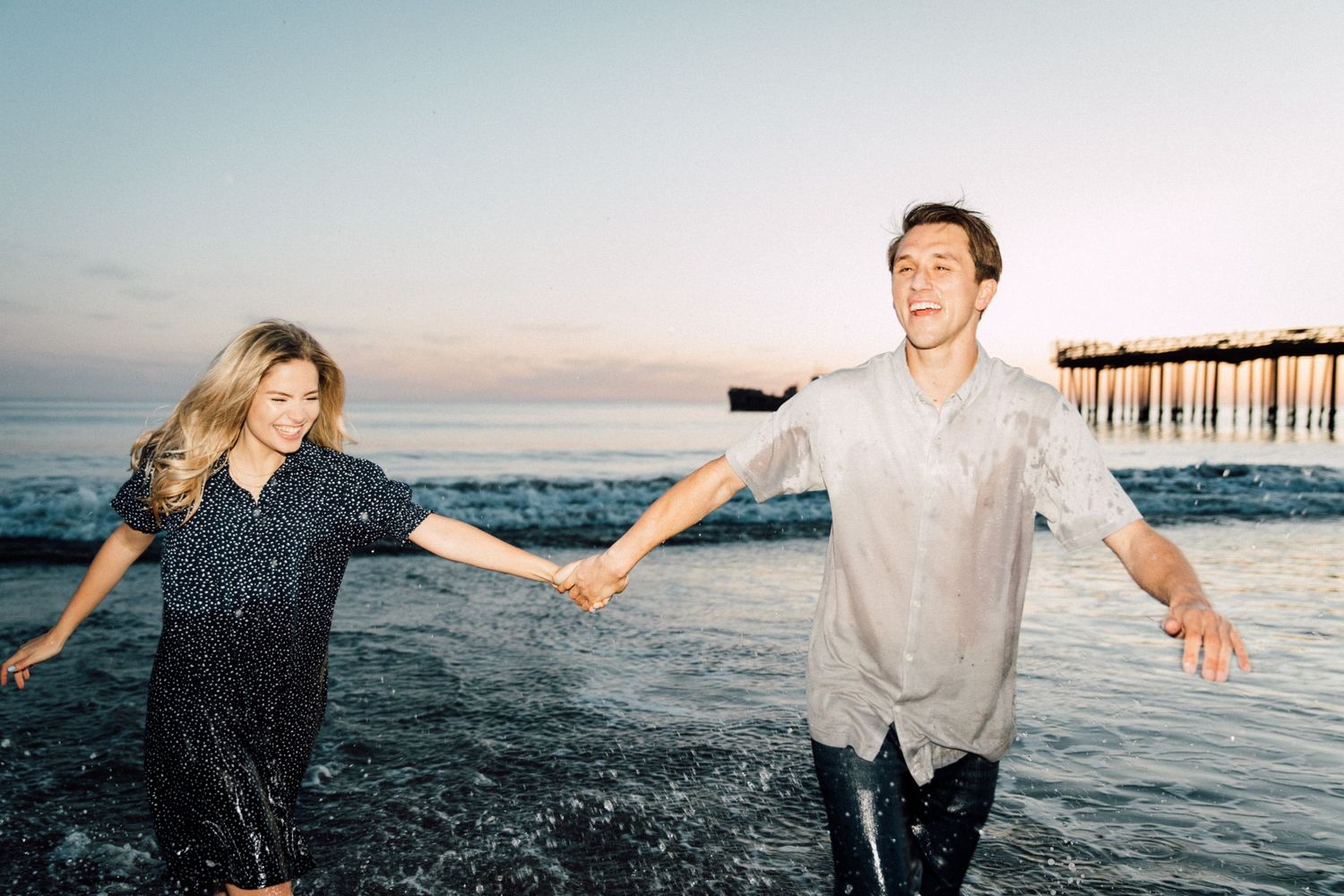 Carefree couple splashing in the ocean at sunset near a pier.