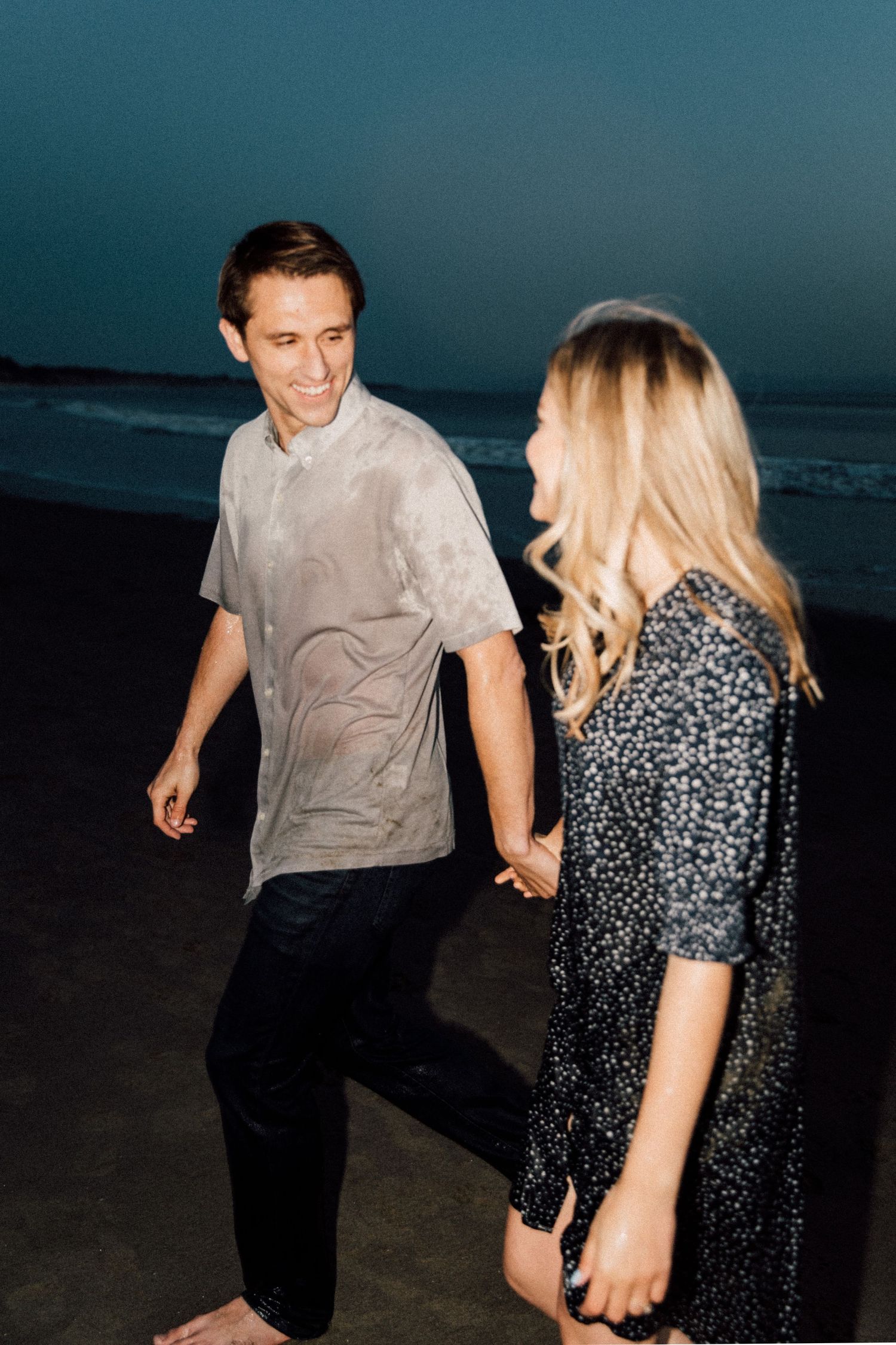 A couple holds hands while walking on a dark beach at dusk, with the woman wearing a patterned dress.