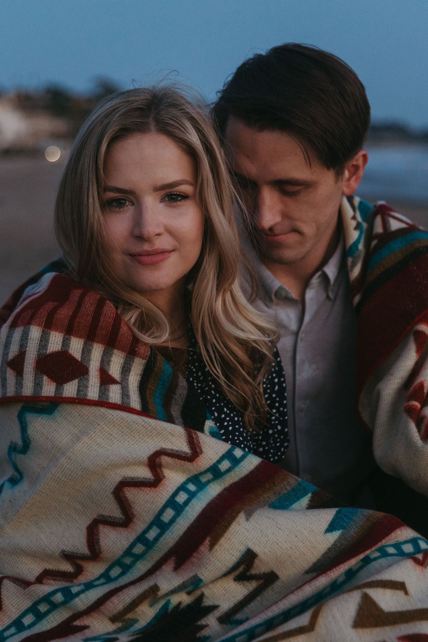A romantic couple wrapped in a southwestern patterned blanket cuddling on the beach at twilight.
