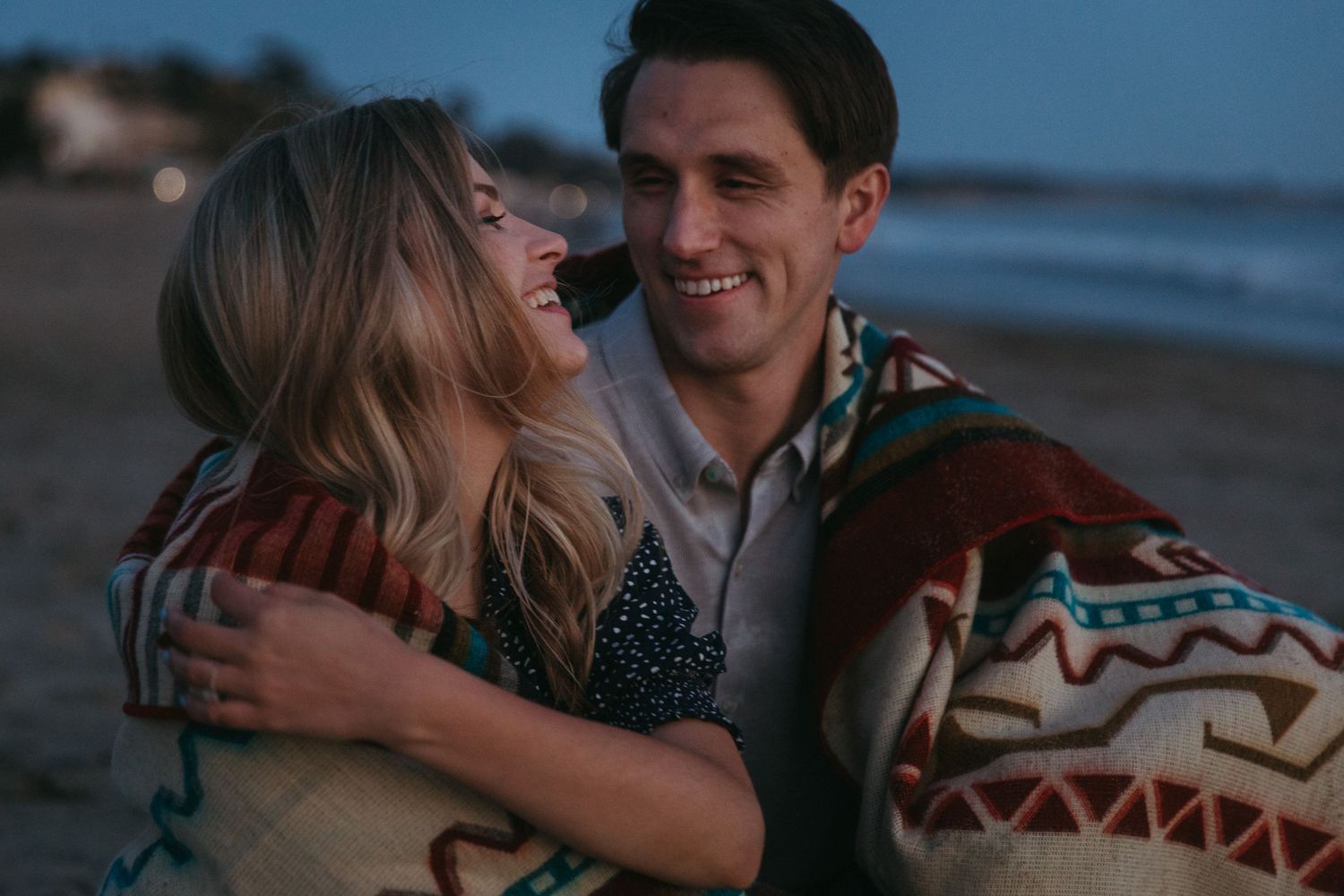 A couple shares a joyful embrace while wrapped in a Native American style blanket on the beach.