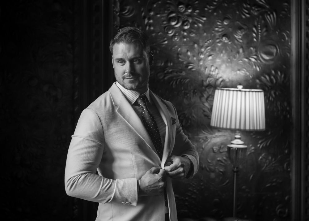 Moody black and white portrait of a groom buttoning up his jacket with a lamp glowing in the background.