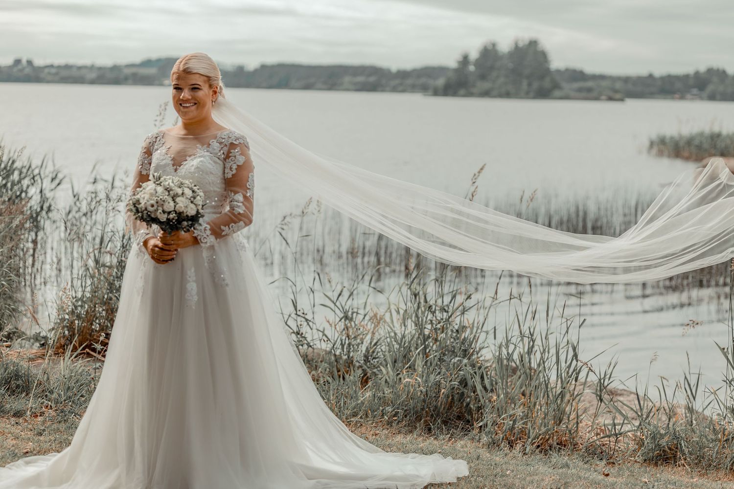 Bride in white lace wedding dress with long flowing veil standing by a serene lake.