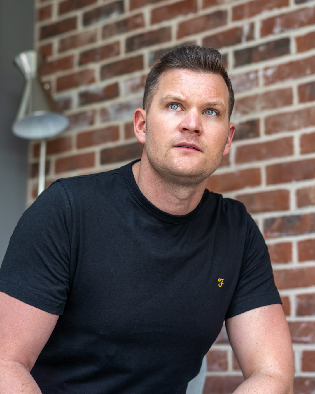Portrait against brick wall featuring someone in a black t-shirt with small logo.