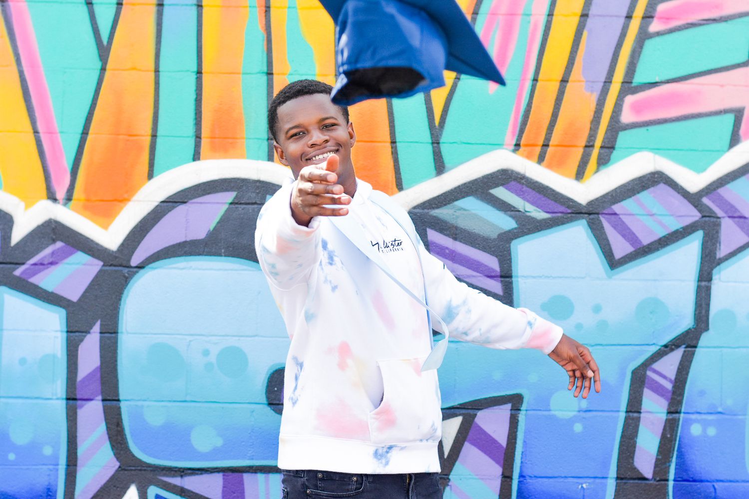 Graduate throws cap in front of colorful graffiti wall during celebration photoshoot.