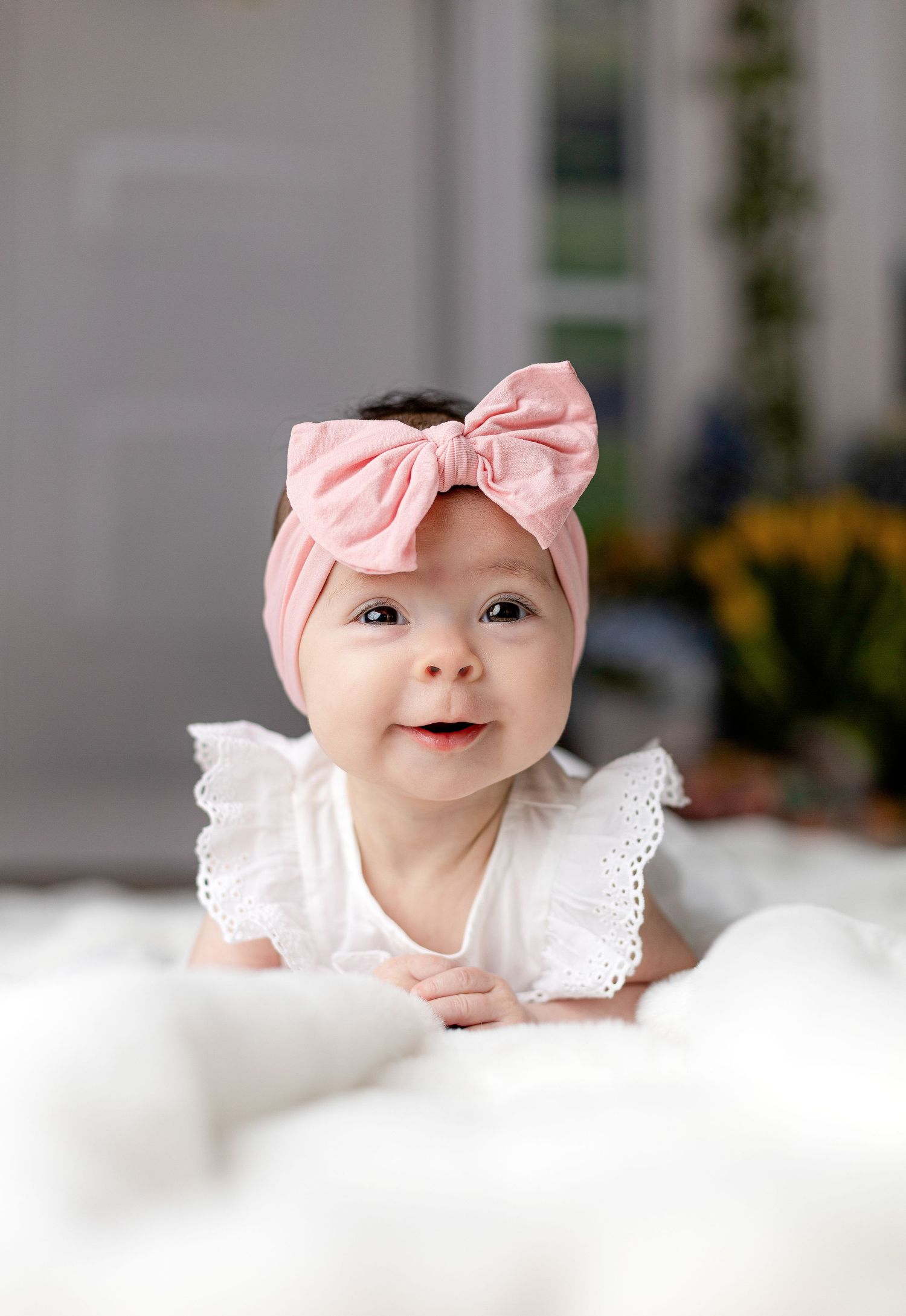 Baby in pink bow headband and white lace outfit lying on fluffy white backdrop.