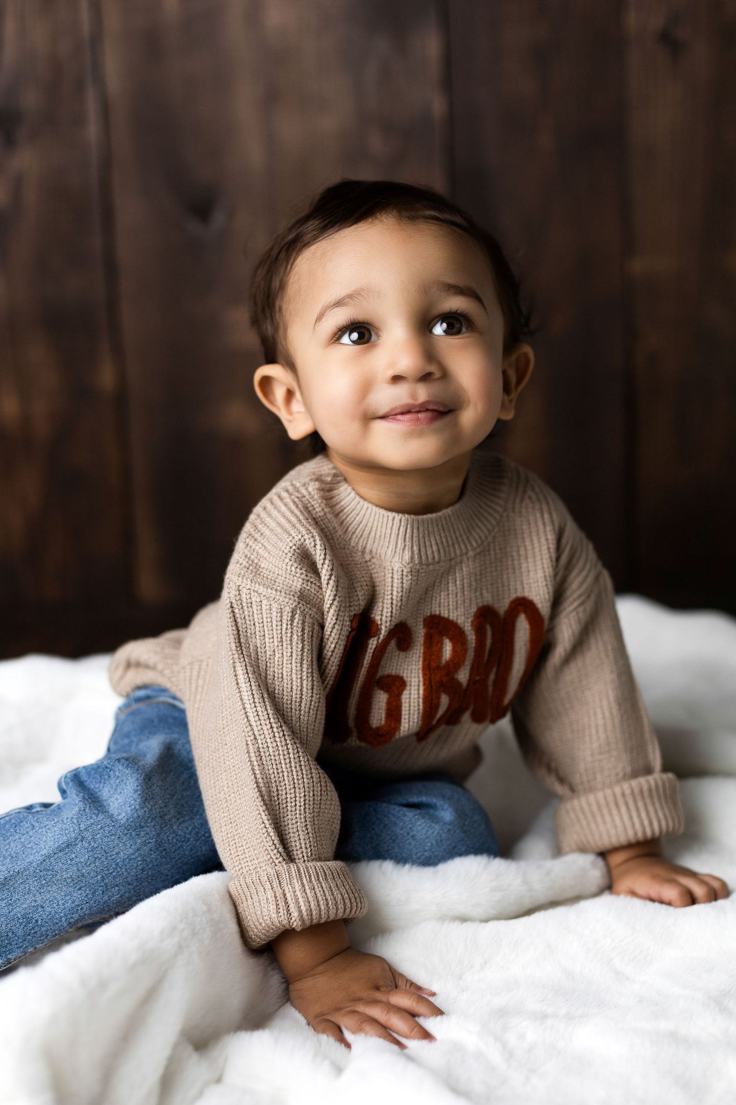 Baby wearing beige sweater lying on white bedding in studio portraits with dark wood background.
