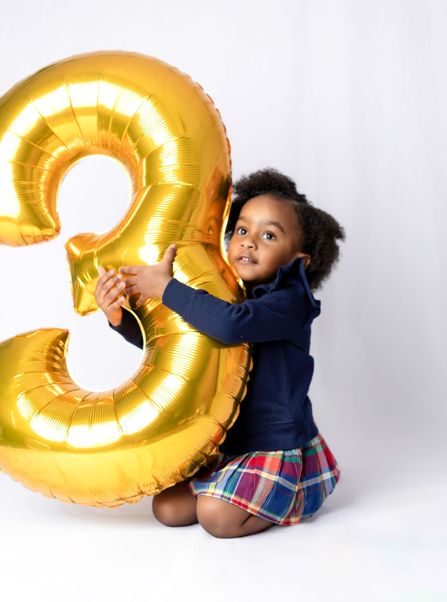 Child playing with large golden number 3 balloon against white background.