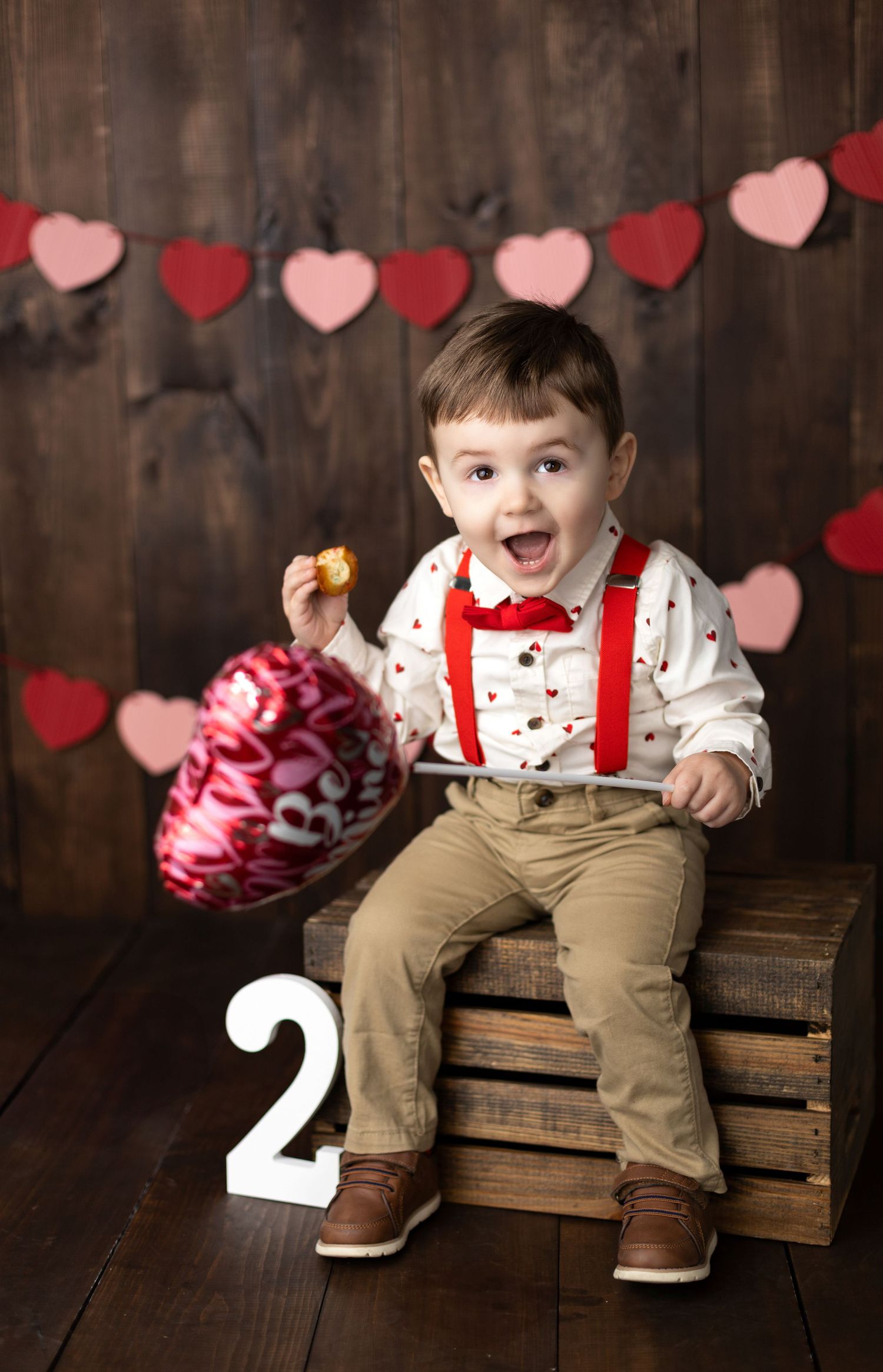 Valentine's themed photo of toddler in suspenders sitting on wooden crates with number two.