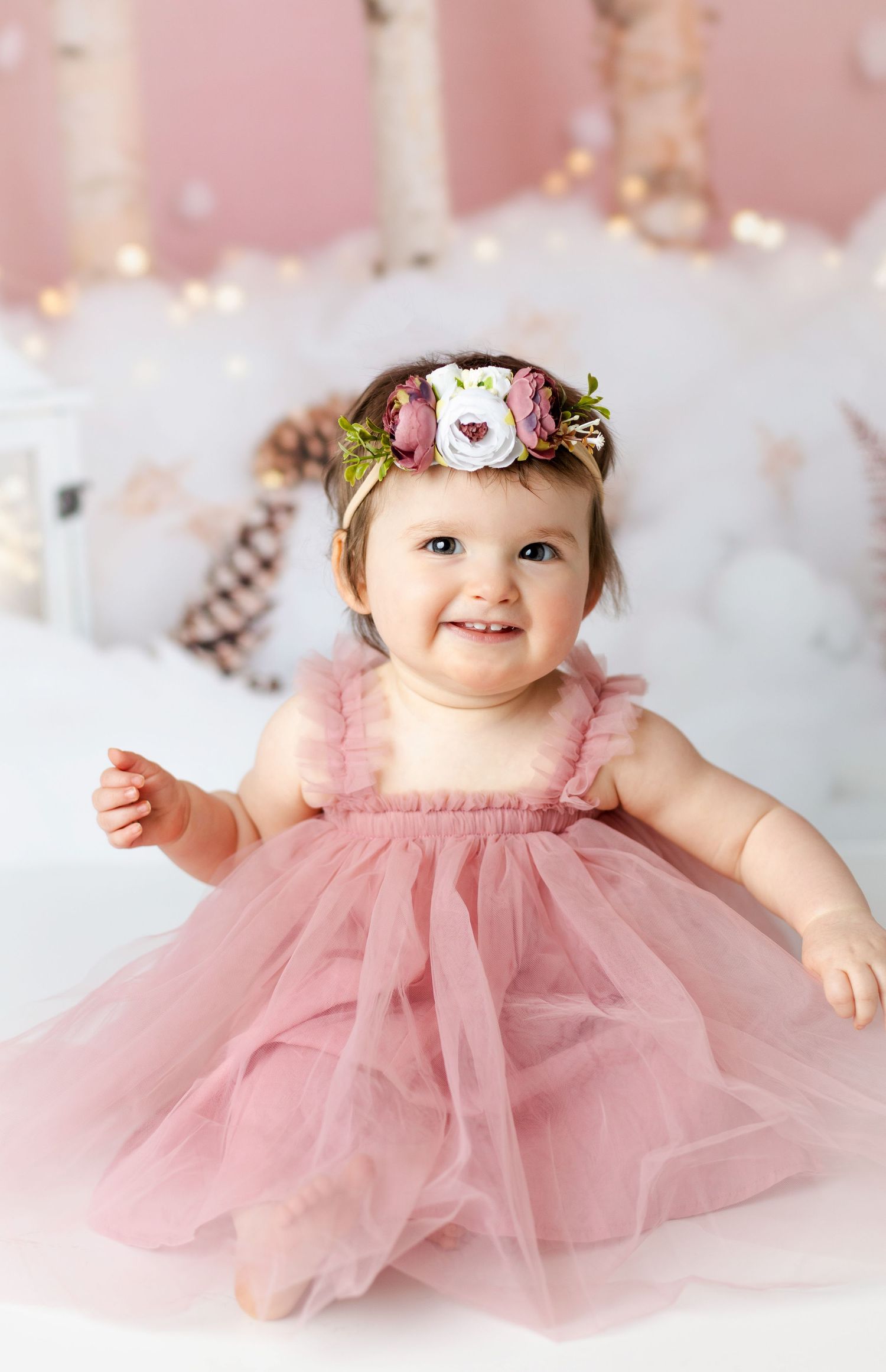 Baby wearing pink tulle dress and floral headband lying on white fluffy backdrop in studio portraits.