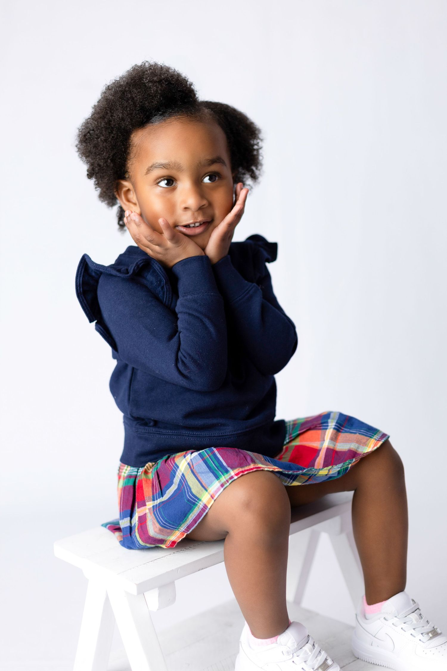 Child in navy sweater and plaid shorts sitting on white platform.