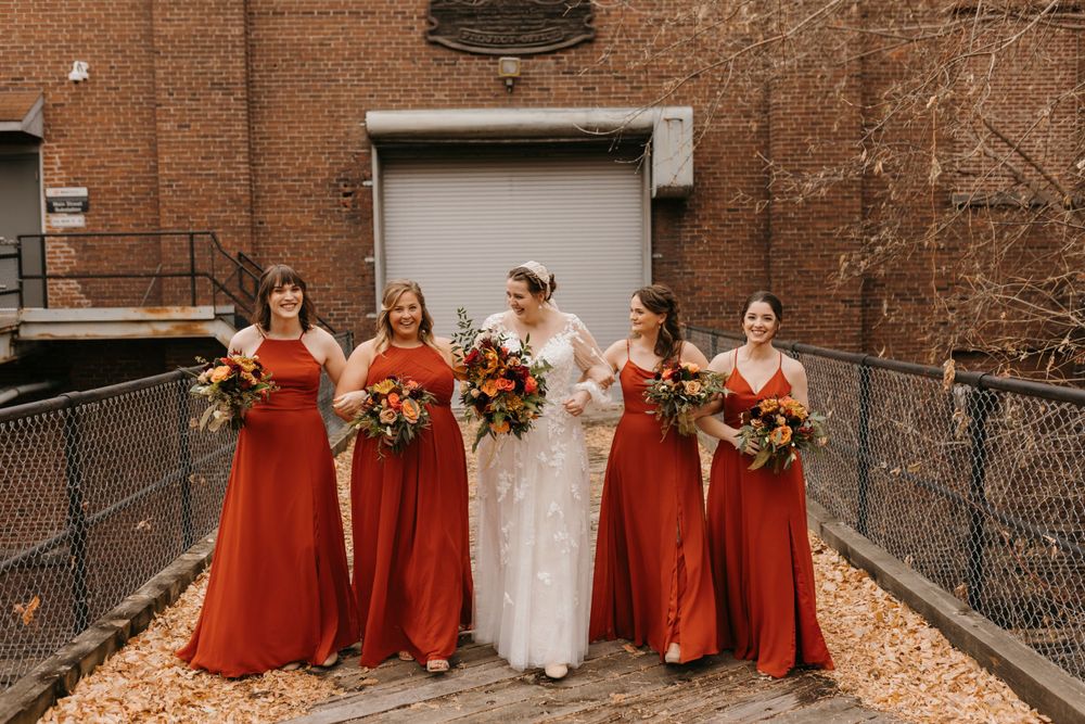 Bride in white lace dress walking with bridesmaids in rust dresses on leaf-covered pathway in fall.