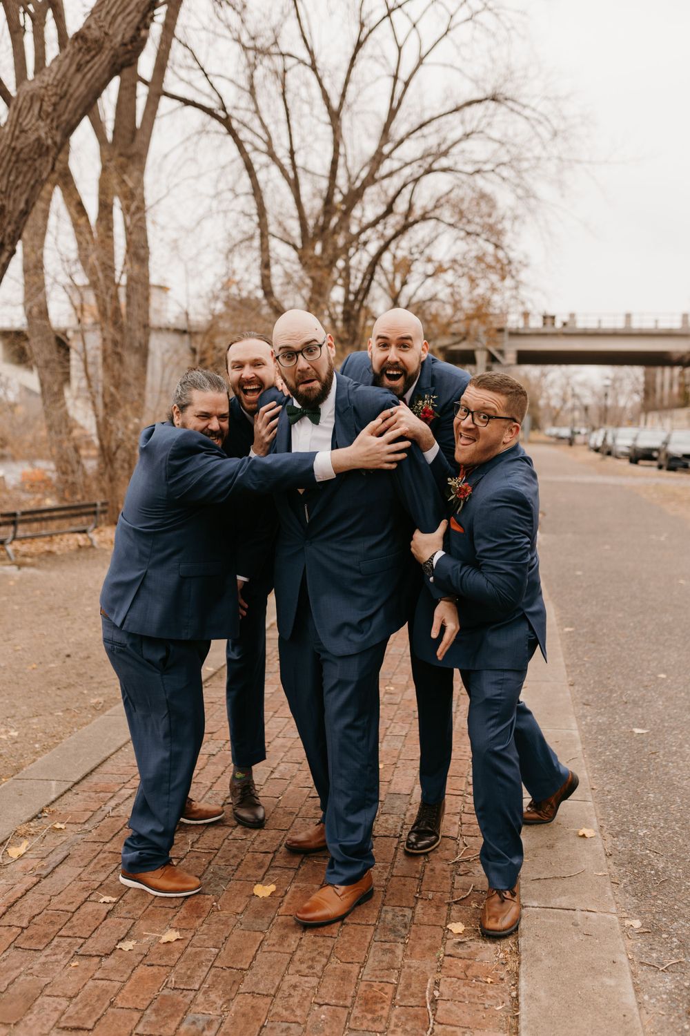 Five men in navy wedding suits laughing and hugging on a brick walkway.