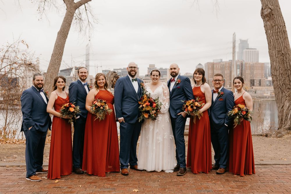Wedding party posing by riverside, bridesmaids in rust-red gowns and groomsmen in navy suits holding fall bouquets.