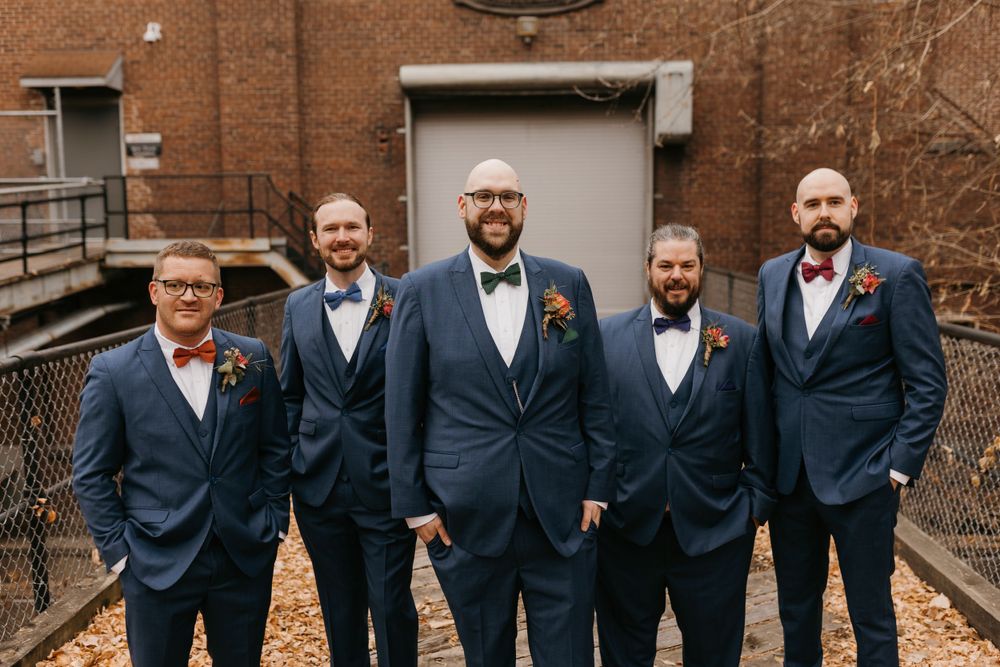 Groom and four groomsmen in navy suits posing formally outside brick building.