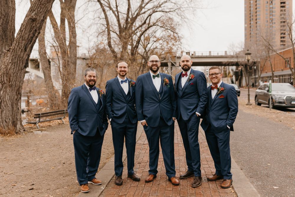 Five groomsmen in navy suits and bow ties smiling together outdoors on a brick pathway in fall setting.