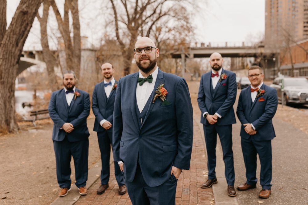 Groom in navy suit with green bow tie stands before four groomsmen outdoors.