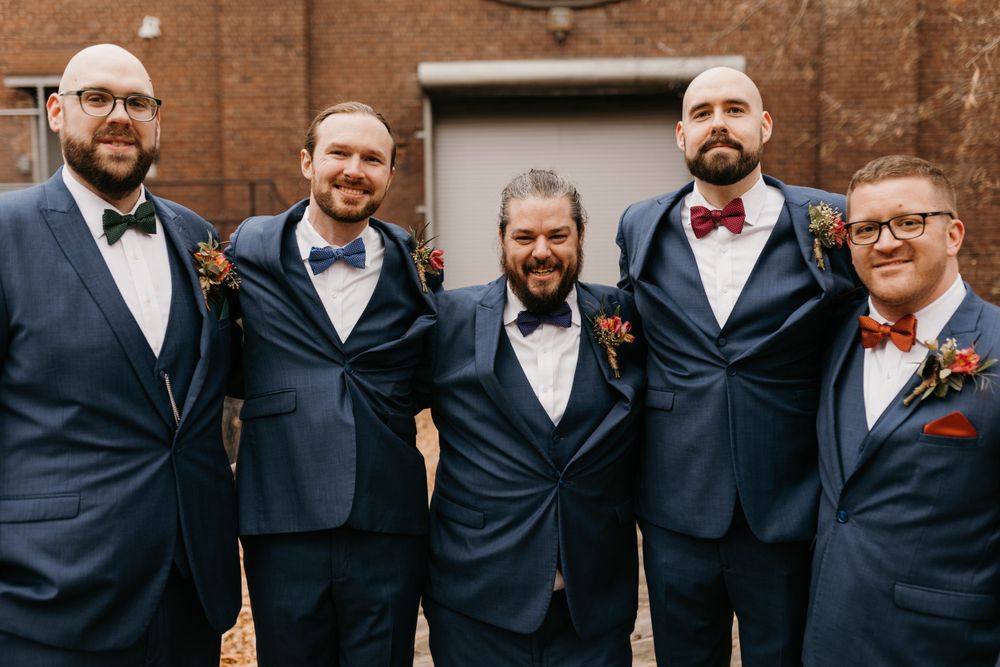 Five groomsmen in navy suits smiling together outside a brick wall venue.