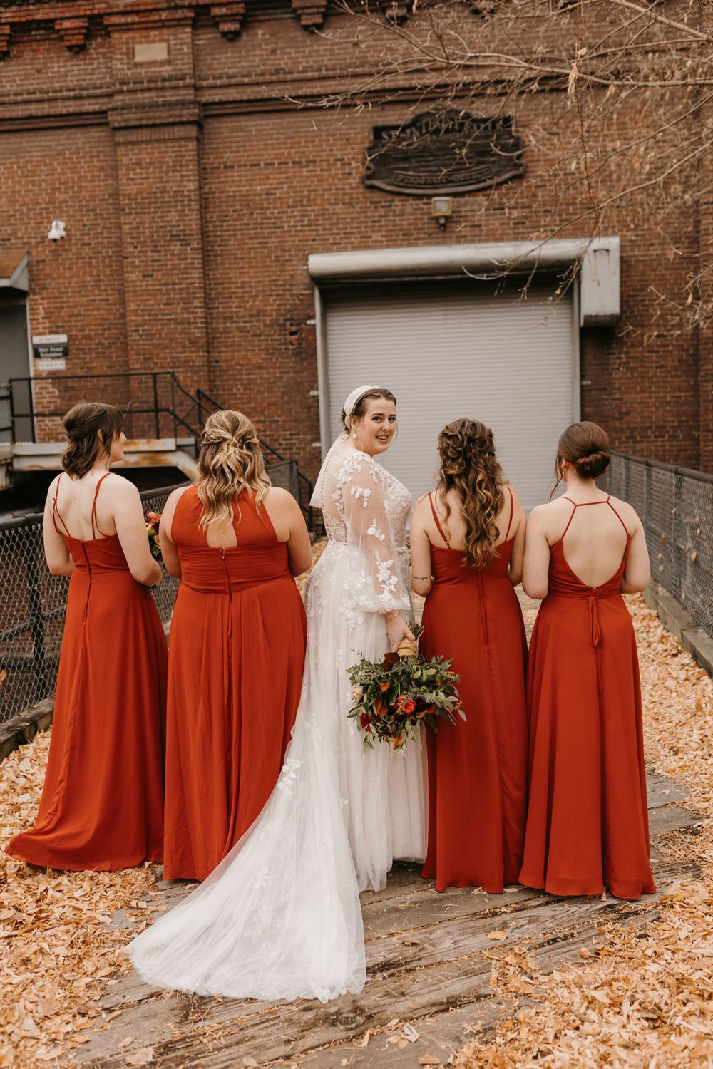 Bride turned showing lace back detail surrounded by four bridesmaids in rust dresses with bouquets.