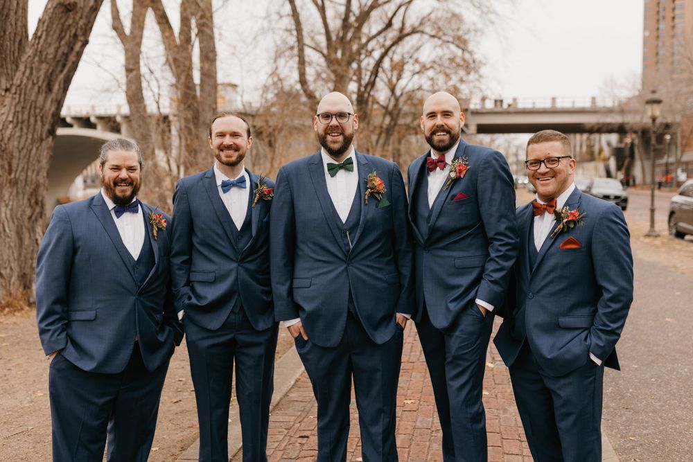 Groom with groomsmen in navy suits and boutonnieres smiling outdoors on a cloudy autumn wedding day.