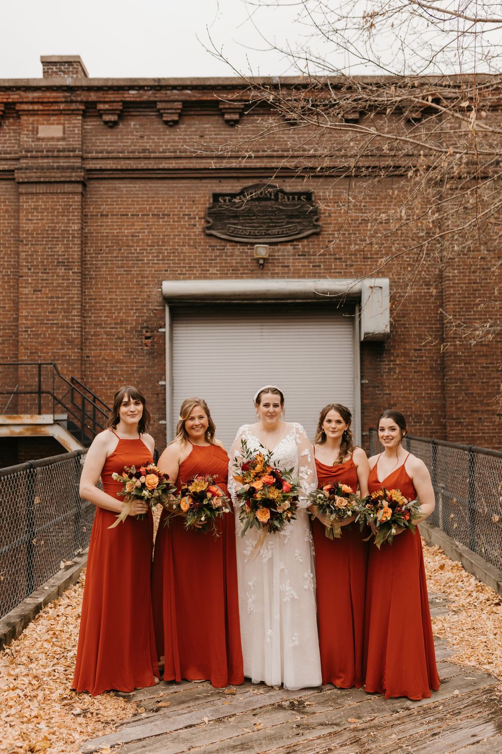 Bridal party of four bridesmaids in burnt orange gowns posing with bride before brick building in fall.