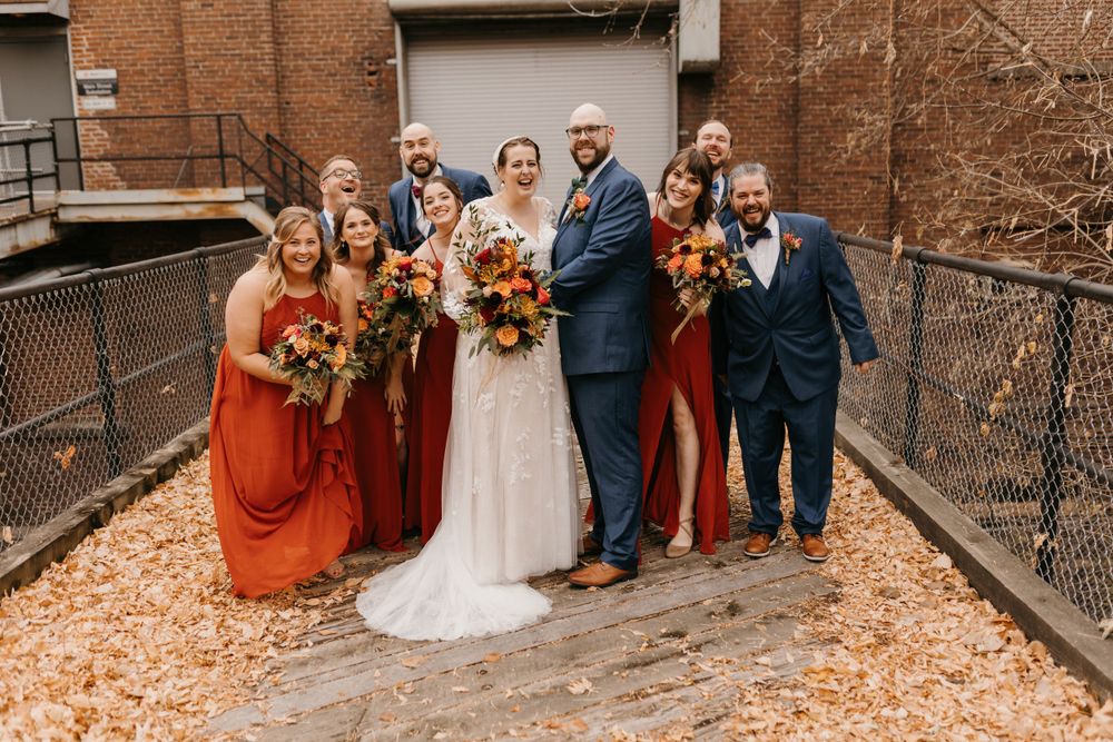 Full wedding party portrait with bride, groom, bridesmaids in orange, and groomsmen in navy on brick path.