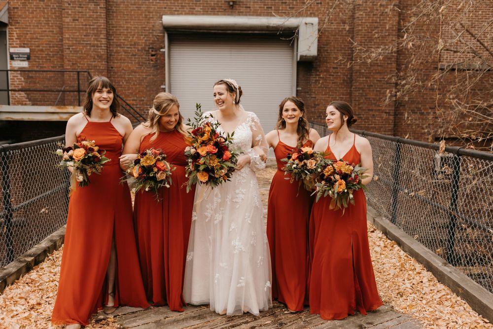 Smiling bride with four bridesmaids in terracotta dresses holding colorful autumn wedding bouquets.
