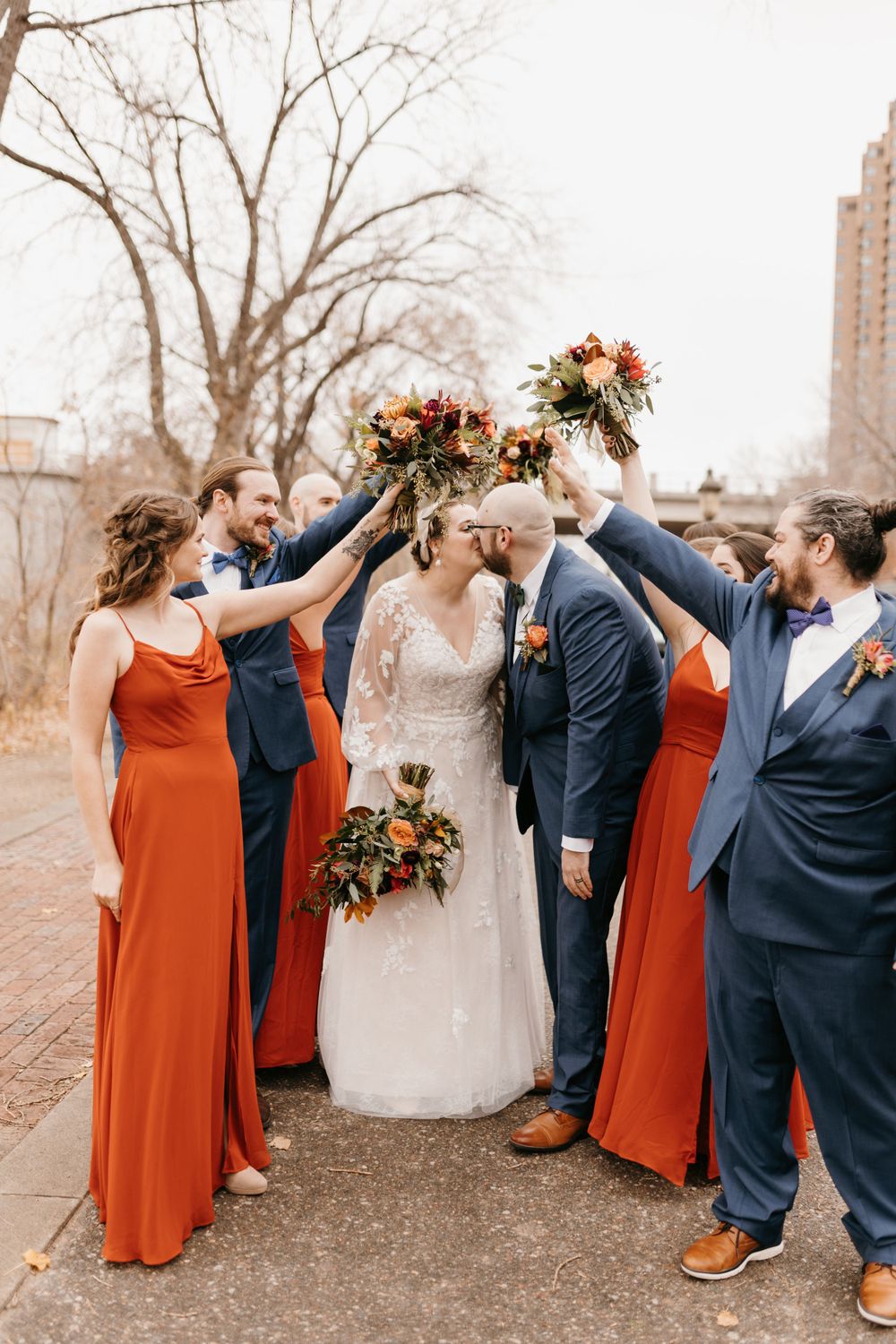 Bride and groom sharing a kiss under raised fall bouquets while wedding party in red and navy surrounds them.