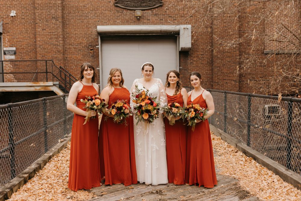 Bride in white lace gown with four bridesmaids in rust orange dresses holding autumn bouquets outdoors.