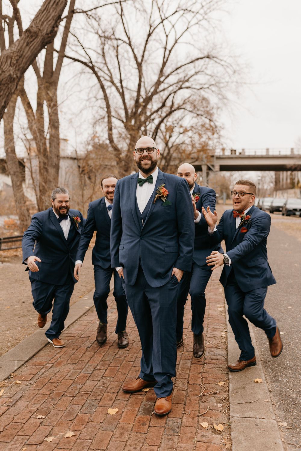 Groom walks confidently on brick path as groomsmen playfully chase behind him.