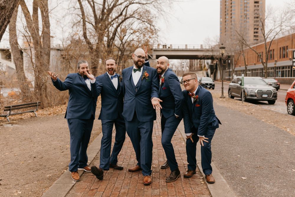 Four groomsmen in navy suits posing playfully on a brick path outdoors.