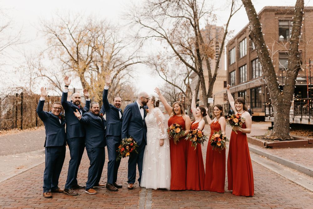 Wedding party celebrating on brick street, bridesmaids in red gowns and groomsmen in navy suits cheering with arms raised.