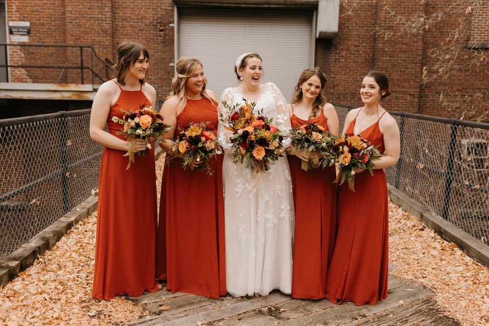 Bride and bridesmaids in red-orange dresses laughing together holding vibrant fall floral bouquets.