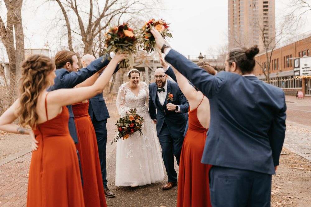 Bride and groom walking under arch of raised fall bouquets held by wedding party in red and navy attire.