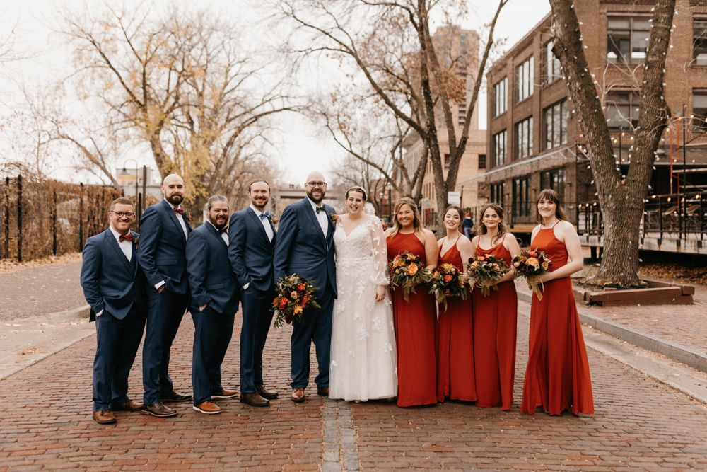 Wedding party poses on brick street in navy suits and red bridesmaid dresses with bouquets.