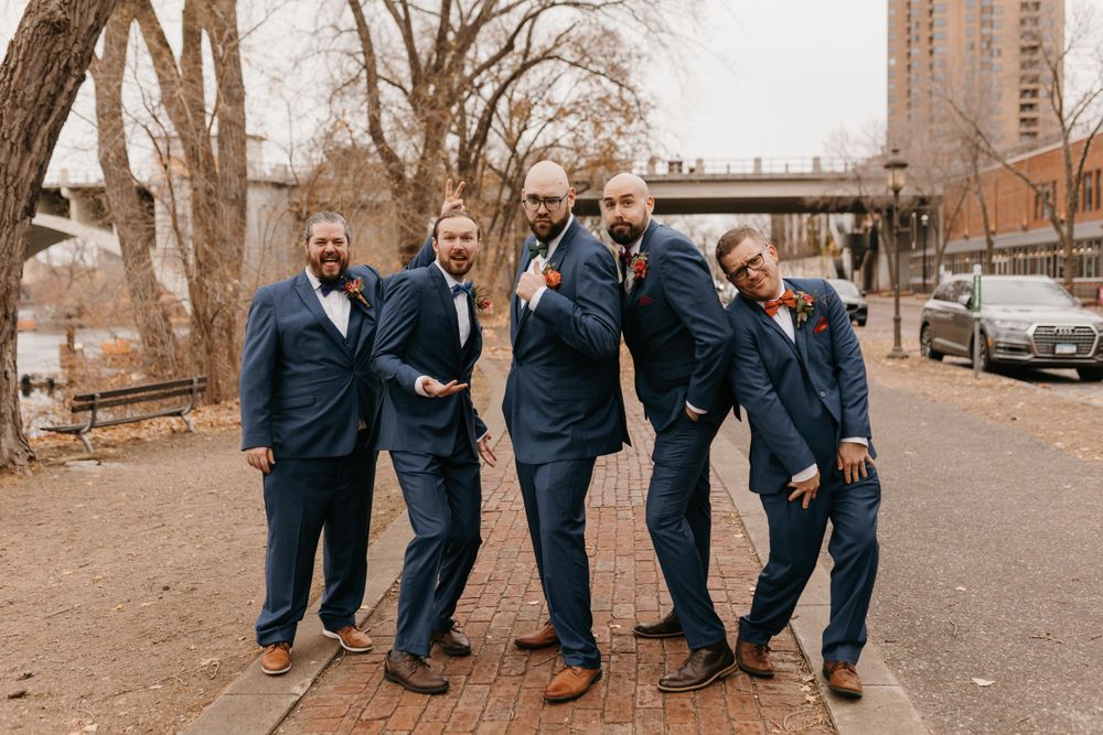 Groomsmen in navy suits striking fun playful poses together on brick pathway during outdoor wedding photos.