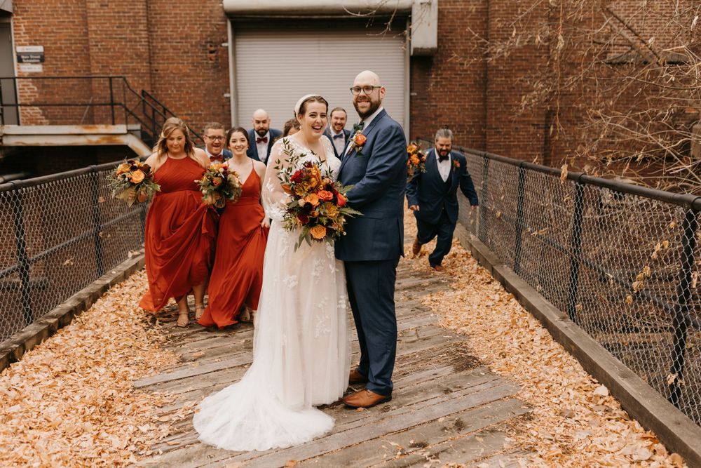 Wedding party group photo with bridesmaids in rust orange and groomsmen in navy suits in fall setting.