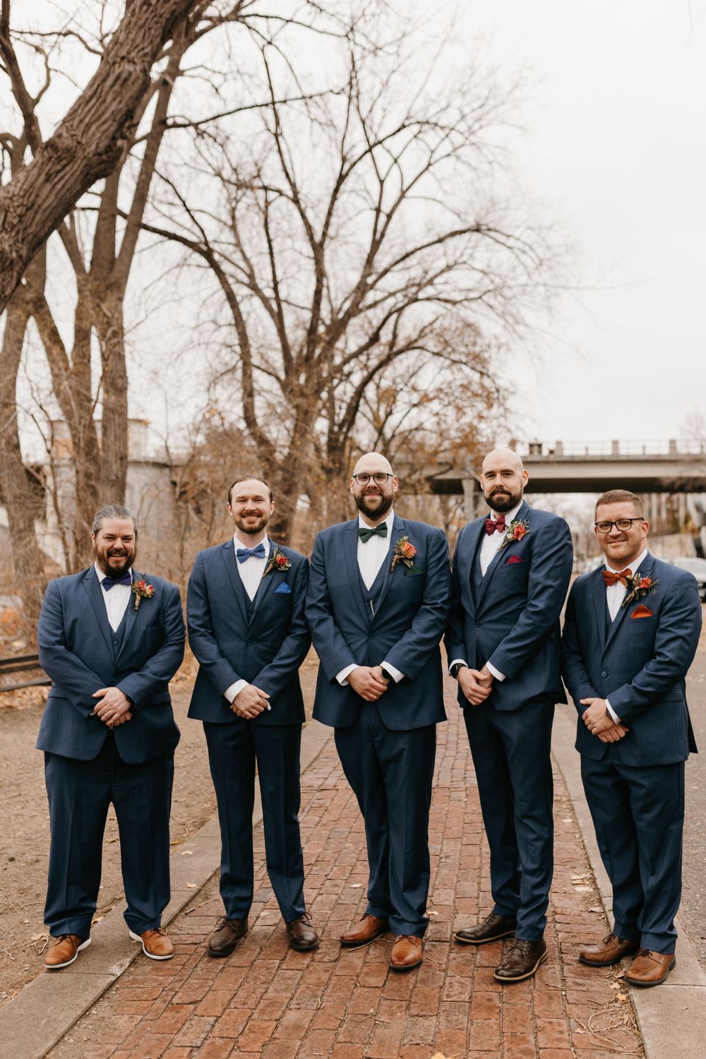Five groomsmen in navy suits standing formally on brick path with bare trees and bridge in background.