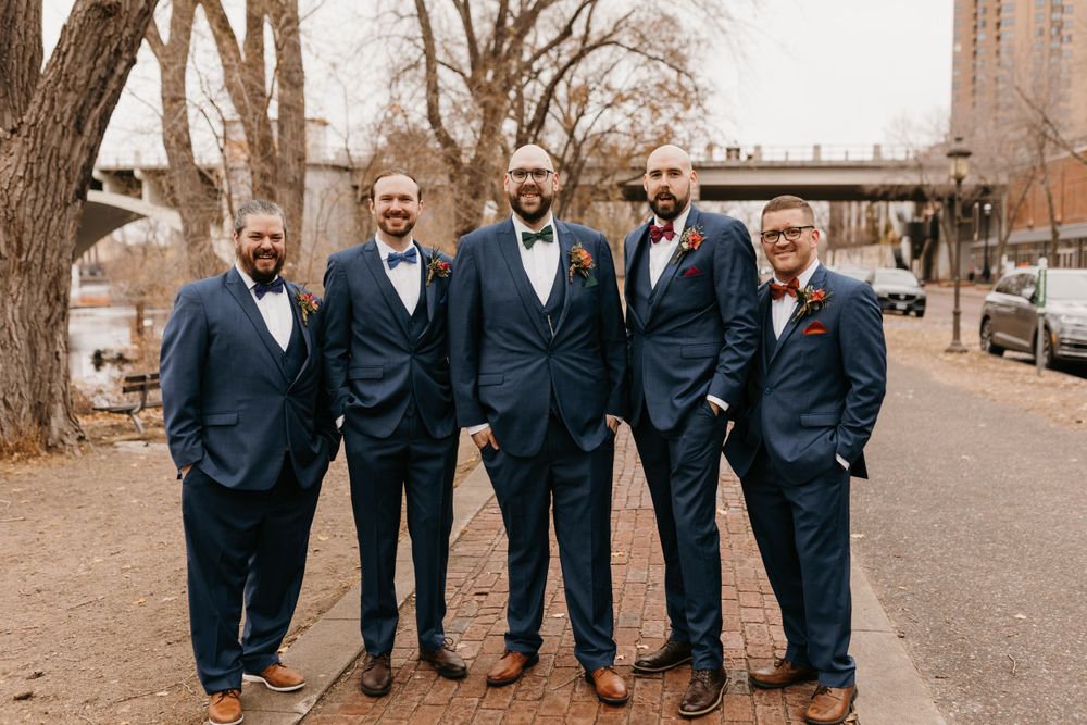 Groom and four groomsmen in matching navy suits laughing together outdoors near riverside on wedding day.