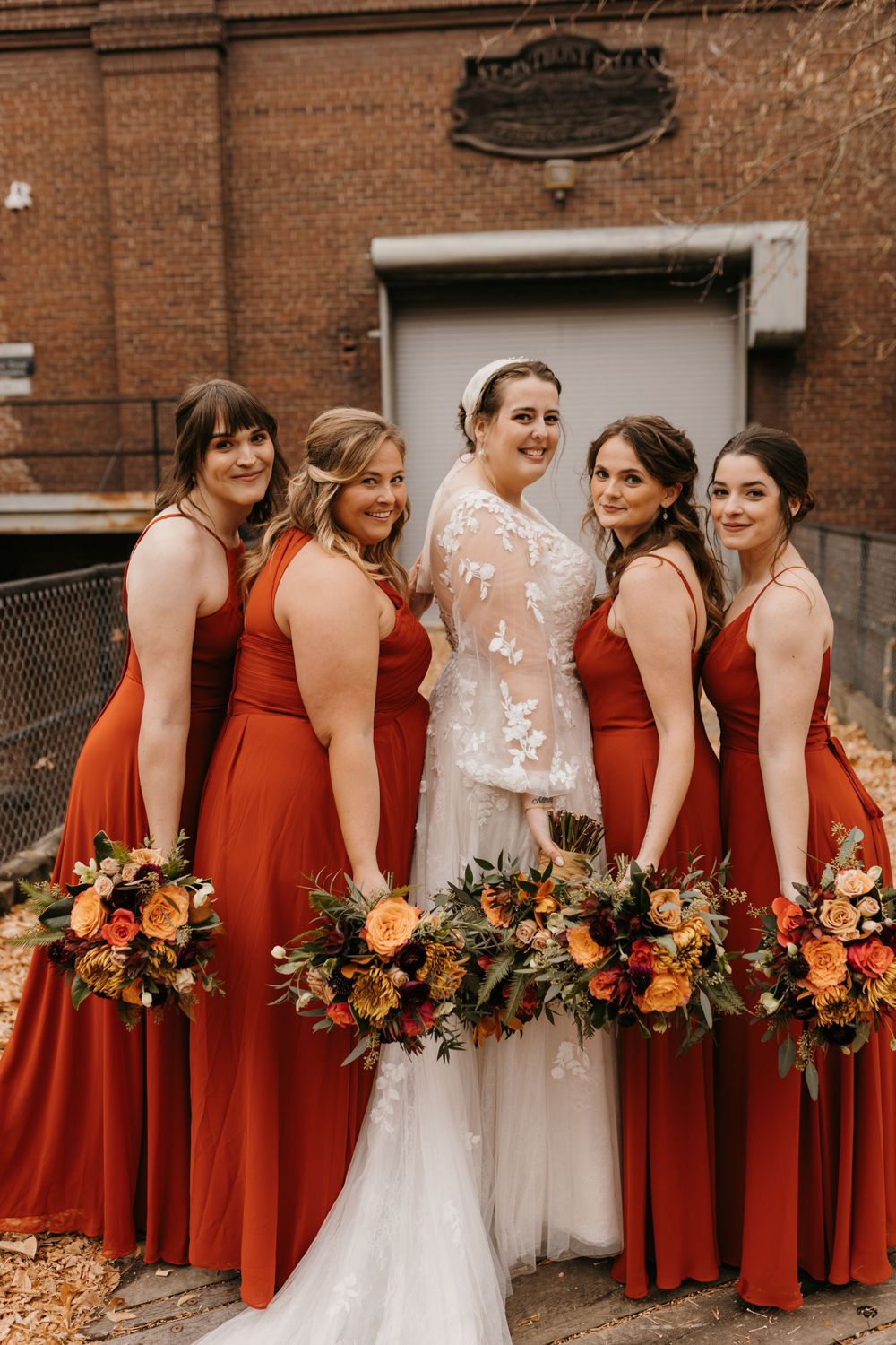 Bride and groom leading wedding party down leaf-covered walkway beside brick building in autumn.