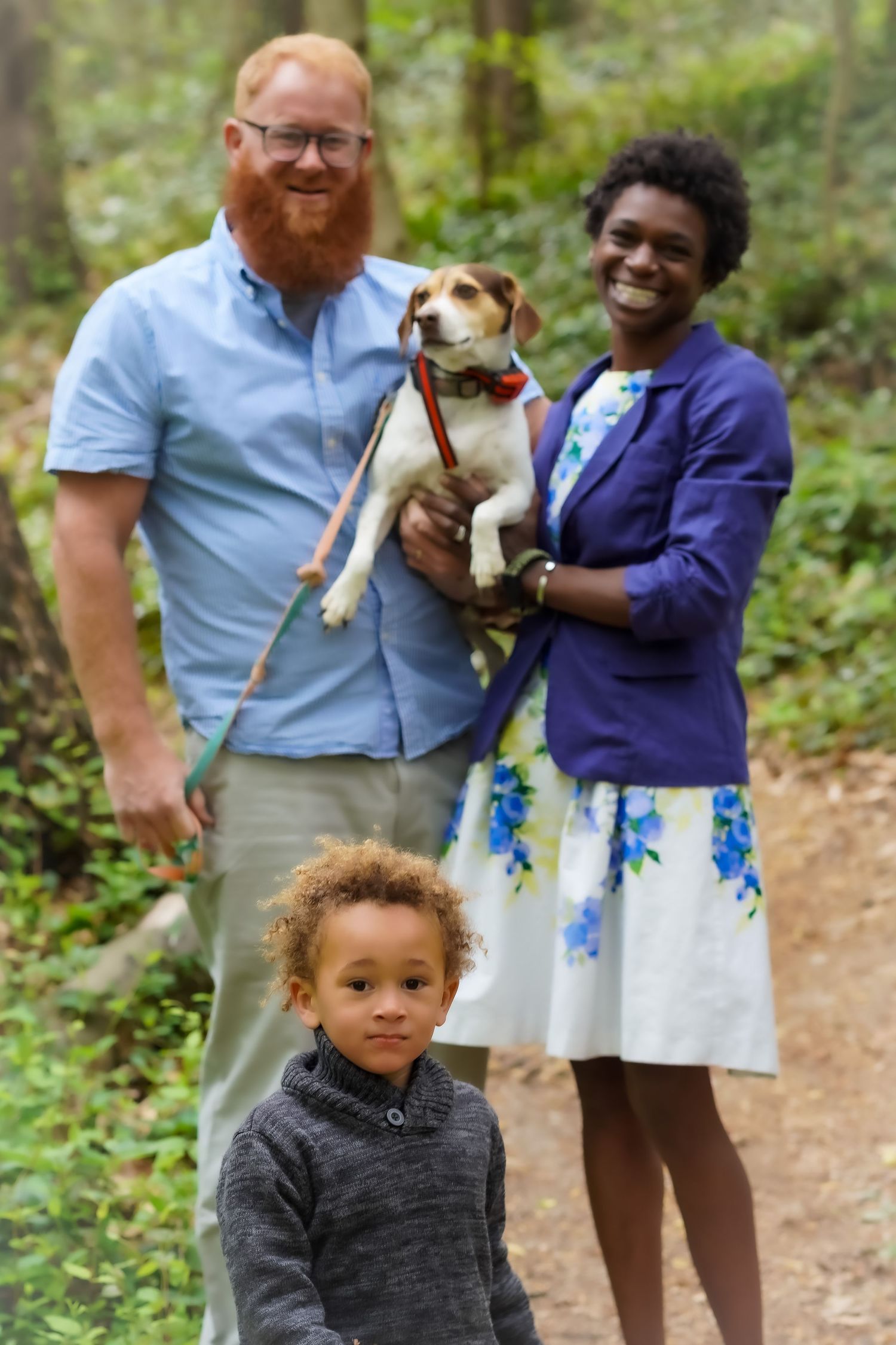A family with their small dog poses together on a forest trail.