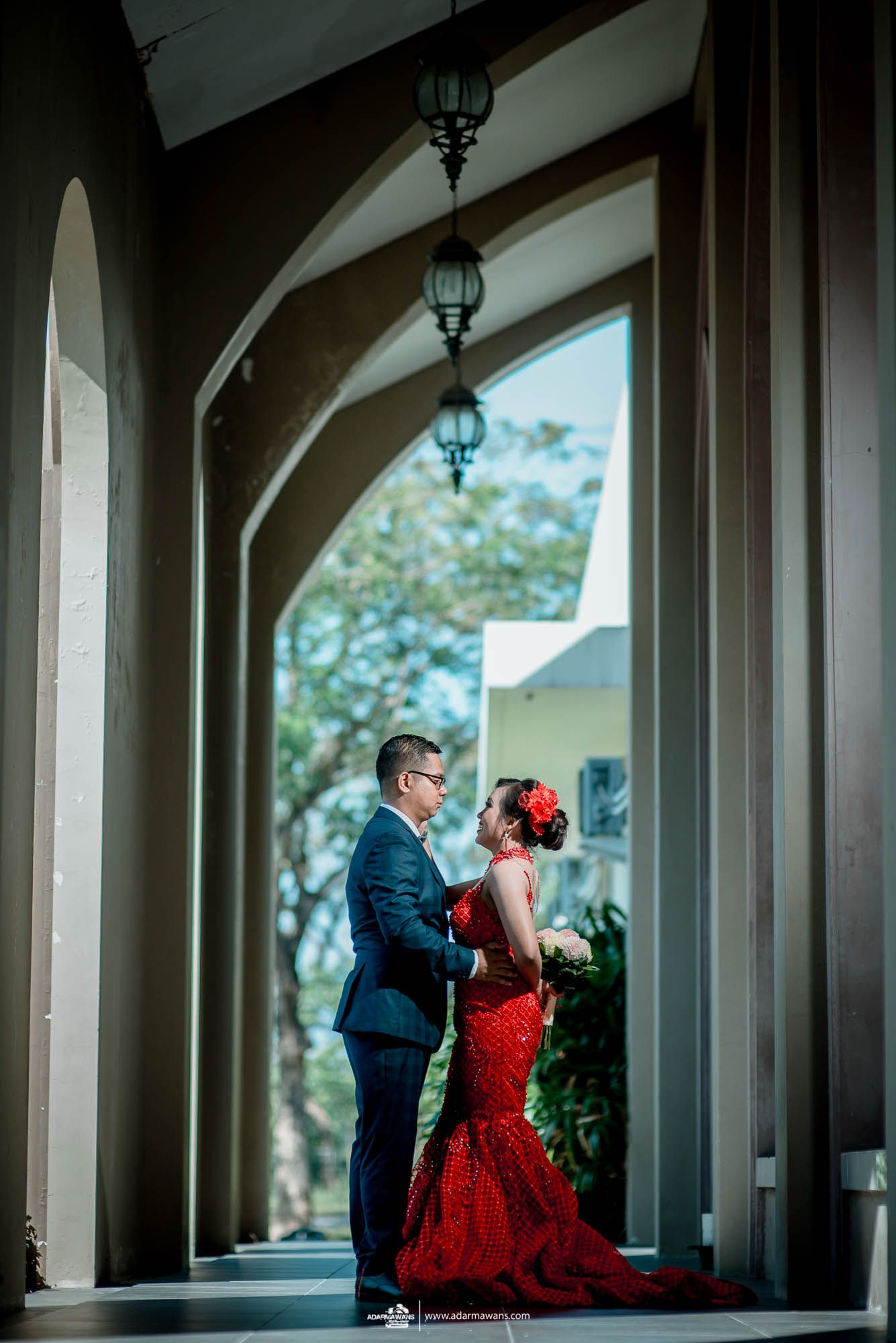 Stylish couple in formal wear standing in architectural archway with hanging lanterns.